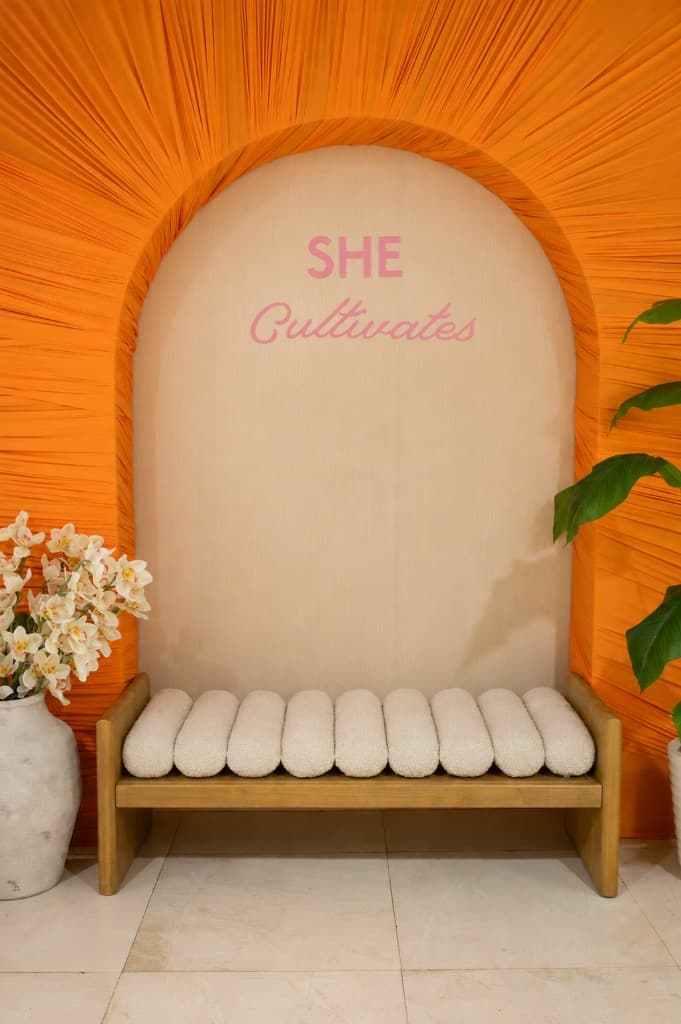 She Cultivates photo backdrop with orange sunburst draping, pink lettering, and a white bouclé bench