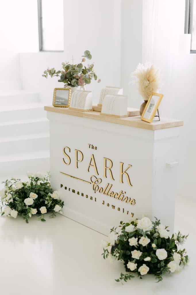 The Spark Collective permanent jewelry station with gold branding, chain displays, and white florals
