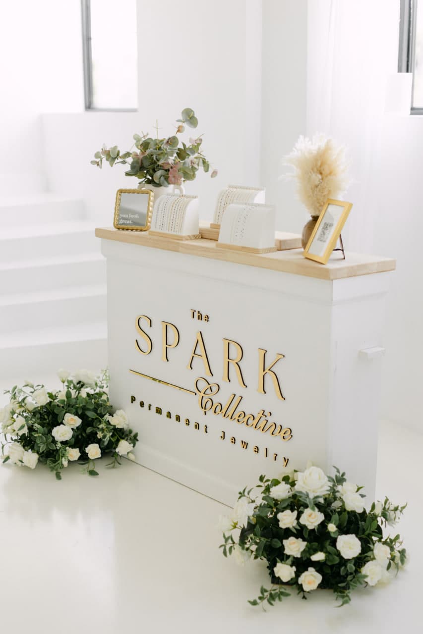 The Spark Collective permanent jewelry station with gold branding, chain displays, and white florals